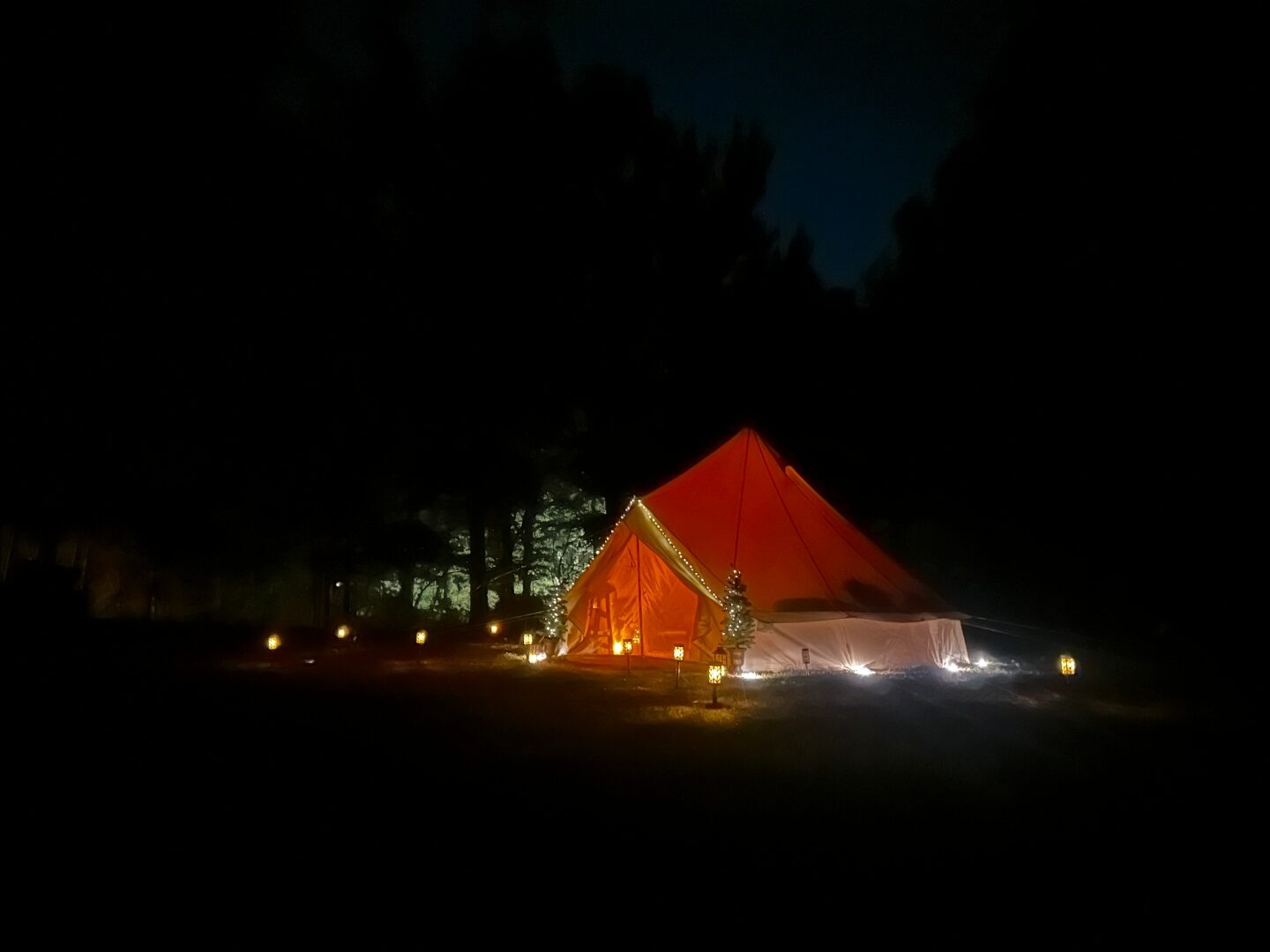Bell tent at night in a forest clearing with warm lanterns glowing inside and out.
